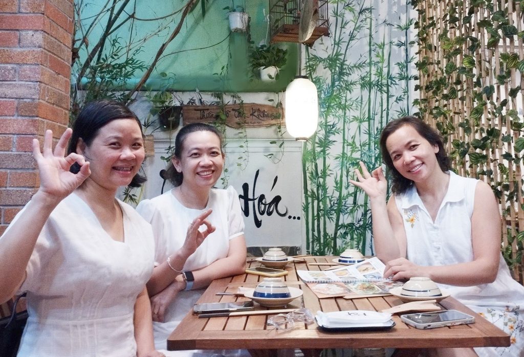 Three women in white attire radiate bright smiles in the serene ambiance of Phuong Mai Vegan Restaurant